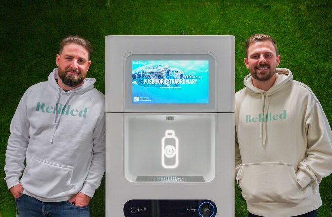 A photo of two men standing with a Refiller drink dispenser between them, against a green background